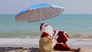 Noel Baba bir plaj şemsiyesinin altında denizin arka planında oturur ve sıcak bir yaz gününün tadını çıkarır. Sıcak ülkelerde Noel ya da yeni yıl tatili. Noel Baba 'nın yaz tatili.