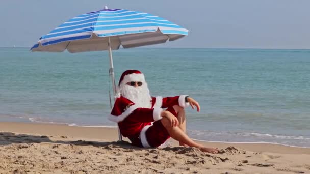 圣诞老人坐在大海背景下的海滩伞下 享受炎热的夏日 在炎热的国家过圣诞节或新年 圣诞老人的暑假 — 图库视频影像
