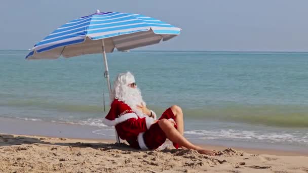 圣诞老人坐在大海背景下的海滩伞下 享受炎热的夏日 在炎热的国家过圣诞节或新年 圣诞老人的暑假 — 图库视频影像