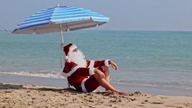 Noel Baba bir plaj şemsiyesinin altında denizin arka planında oturur ve sıcak bir yaz gününün tadını çıkarır. Sıcak ülkelerde Noel ya da yeni yıl tatili. Noel Baba 'nın yaz tatili.