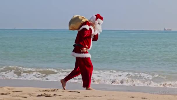 夏天炎热的一天 圣诞老人背着一袋礼物沿着沙滩沿着冲浪线散步 在炎热的国家过圣诞节或新年 圣诞老人的暑假 — 图库视频影像