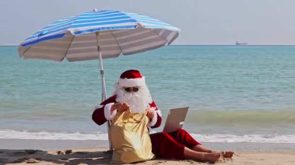 在一个炎热的夏日里 圣诞老人坐在大海的背景下的海滩伞下 在笔记本电脑上输入一些东西 查看一袋礼物 圣诞老人的暑假 — 图库视频影像