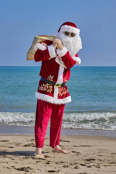 Sıcak bir yaz gününde Noel Baba denizde. Noel Baba denizin önündeki kumlu bir sahilde yalınayak duruyor. Omzunda hediyelerle bir çanta tutuyor ve elini sallıyor..