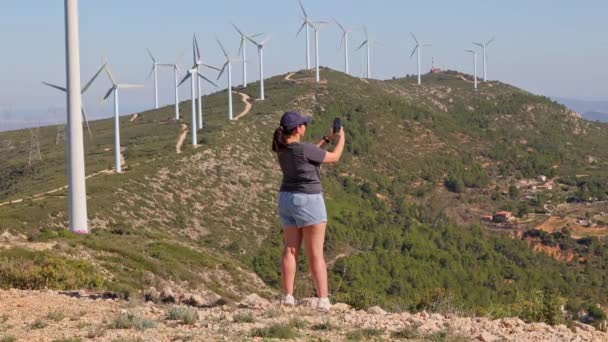 ランドスケープビデオを撮影する少女 スマートフォンでソーシャルメディアの写真 女の子は彼の携帯電話を使用して山の風力タービンの水平パノラマを取ります — ストック動画