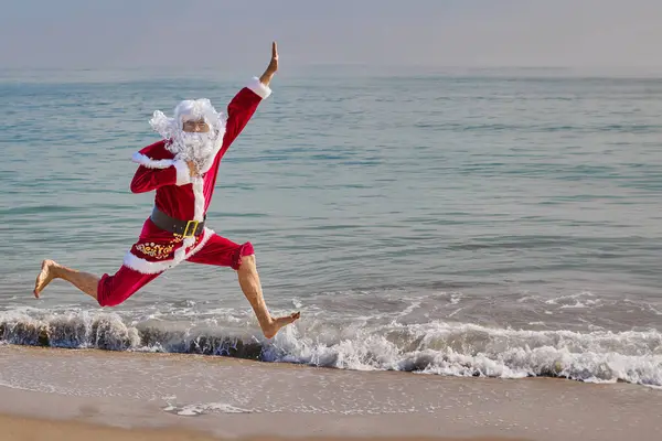 Noel Baba sıcak bir yaz gününde sahile, denizin arka planına atlıyor. Sıcak ülkelerde Noel ya da yeni yıl tatili. Noel Babalar yaz tatili.