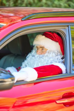 Gözlüklü ve beyaz eldivenli Noel Baba kırmızı bir arabada oturur ve önüne bakar.