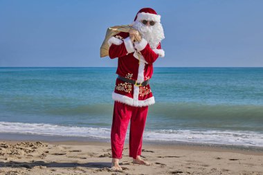 Sıcak bir yaz gününde Noel Baba denizde. Noel Baba denizin önündeki kumlu bir sahilde yalınayak duruyor. Omzunda hediyelerle bir çanta tutuyor ve elini sallıyor..