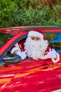 Güneş gözlüklü ve beyaz eldivenli Noel Baba kafasını kırmızı bir arabanın camından dışarı çıkarıyor ve elleriyle bir jest yapıyor. Yeşil Orman geçmişi. Yakın plan portre