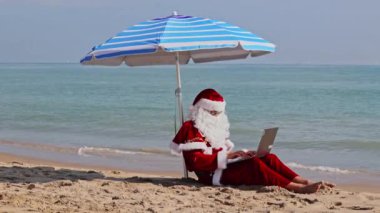 Noel Baba sıcak bir yaz gününde plaj şemsiyesinin altında denizin arka planında oturur ve dizüstü bilgisayarına bir şeyler yazar. Sıcak ülkelerde Noel ya da yeni yıl tatili. Noel Baba 'nın yaz tatili.