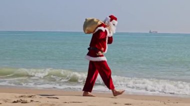 Omzunda hediyelerle dolu bir çantayla Noel Baba sıcak bir yaz gününde dalgalar boyunca kumlu sahil boyunca yürür. Sıcak ülkelerde Noel ya da yeni yıl tatili. Noel Baba 'nın yaz tatili.