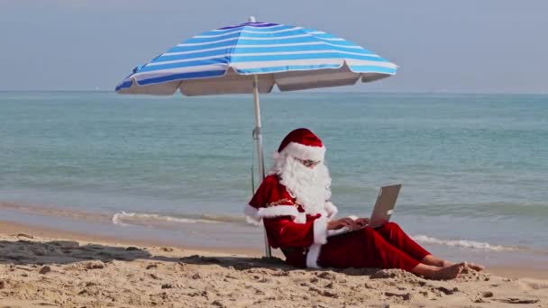 在炎热的夏日 圣诞老人坐在海边的伞下 靠着大海 在笔记本电脑上打字 在炎热的国家过圣诞节或新年 圣诞老人的暑假 — 图库视频影像