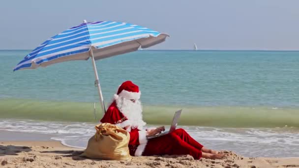 在炎热的夏日 圣诞老人坐在海边的伞下 靠着大海 在笔记本电脑上打字 在炎热的国家过圣诞节或新年 圣诞老人的暑假 — 图库视频影像