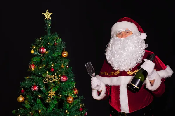 Noel Baba, siyah arka planda güzel dekore edilmiş ve aydınlatılmış bir Noel ağacının yanında duruyor. Elinde boş bir bardak ve elinde bir şişe şampanya tutuyor. Xmas dekorasyon kavramı.