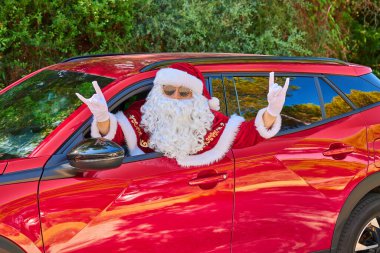 Güneş gözlüklü ve beyaz eldivenli Noel Baba kafasını kırmızı bir arabanın camından dışarı çıkarıyor ve eliyle bir jest yapıyor. Yeşil Orman geçmişi. Yakın plan portre
