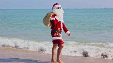 Omzunda hediyelerle dolu bir çantayla Noel Baba sıcak bir yaz gününde dalgalar boyunca kumlu sahil boyunca yürür. Sıcak ülkelerde Noel ya da yeni yıl tatili. Noel Baba 'nın yaz tatili.