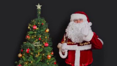 Noel Baba bir bardak köpüklü şarapla Noel ağacının yanında duruyor. Sosyal bir jest gibi. Siyah bir arkaplanda.