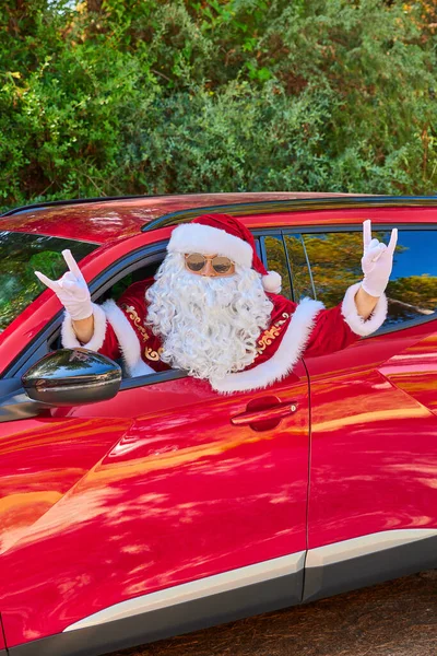 Güneş gözlüklü ve beyaz eldivenli Noel Baba kafasını kırmızı bir arabanın camından dışarı çıkarıyor ve eliyle bir jest yapıyor. Yeşil Orman geçmişi. Yakın plan portre