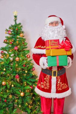 Noel Baba süslenmiş bir Noel ağacının yanında hediyelerle dolu kutular tutuyor. Beyaz arkaplanda.