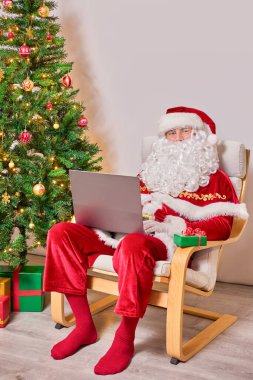 Noel Baba dizüstü bilgisayarı olan bir sandalyede oturuyor. Noel ağacının yanında. Beyaz arkaplanda.