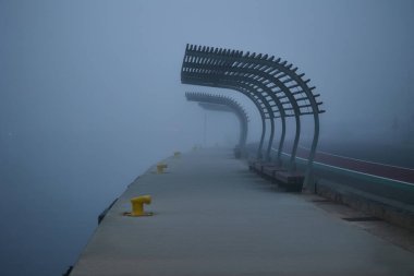 Yoğun sis sırasında boş iskele boyunca banklar. Arka planda belirsiz siluetler.