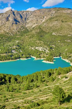 Guadalest rezervuarının manzarası. İspanya 'nın Alicante eyaletinde Guadalest' in doğasında mavi gökyüzüne karşı uzanan yeşil ağaçlarla çevrili sakin bir turkuaz gölü.