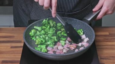 Şef brokoli, pastırma ve ıspanaklı yumurta kızartması. Kızartma tavasında brokoli ve pastırma kızartan bir kadın..