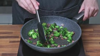 Şef brokoli, pastırma ve ıspanaklı yumurta kızartması. Kızartma tavasında brokoli ve pastırma kızartan bir kadın..