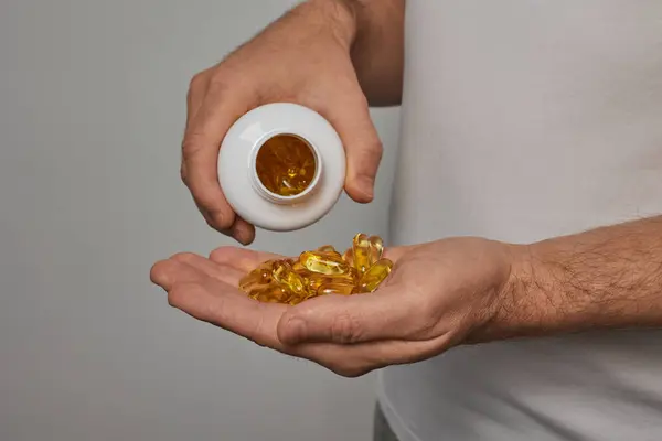Beyaz tişörtlü genç bir adam eline hap döküyor. Omega 3 Altın Balık Yağı Kapsülleri. Beyaz arkaplanda yakın plan.