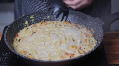 Bir kadın tavada şarap soslu midyeli spagetti pişiriyor. Spatulayla spagettiyi karıştırır..