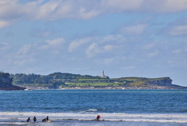 İnsanlar Santander 'deki Cape Mayor deniz fenerinin arka planında Somo sahilinde sörf yapıyor. Somo, İspanya - 13 Eylül 2024.