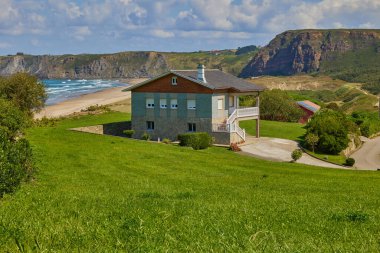Kumlu bir sahile bakan yeşil bir tepede küçük bir otel ve parlak mavi bir gökyüzünün altındaki uçurumlar. San Juan de Nieva, Asturias - 17 Eylül 2024.