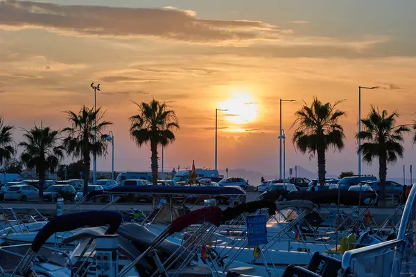 İspanya 'daki Denia Marina' da gün batımı manzarasında, canlı turuncu ve mor gökyüzü altında rıhtımlı tekneler, palmiye ağaçları ve arabalar yer alıyor. Denia, İspanya - 31 Mayıs 2025.
