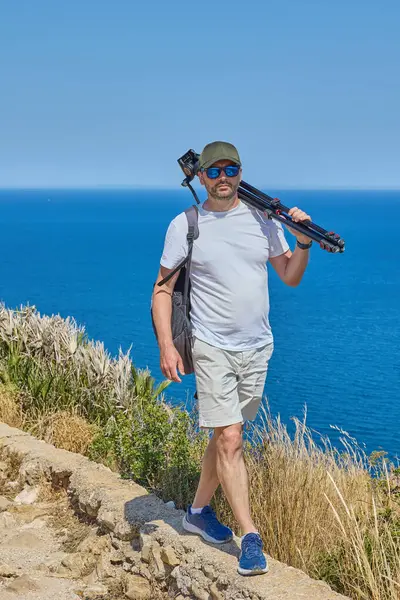 Beyaz tişörtlü, güneş gözlüklü ve şapkalı bir adam Akdeniz 'in üzerindeki sahil yolunda yürüyor. Omzunda ve sırt çantasında bir tripod taşıyor. Bu da fotoğrafçılık ya da video gezisi olduğunu gösteriyor..