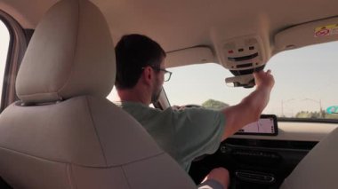 Güneş gözlüklü genç bir adamın güneşli bir günde manzaralı bir kır yolunda araba sürüşünün arka koltuğu.