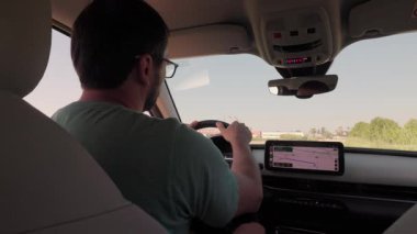 Güneş gözlüklü genç bir adamın güneşli bir günde manzaralı bir kır yolunda araba sürüşünün arka koltuğu.