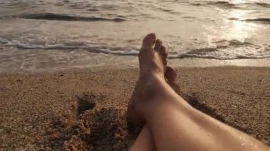 Deniz dalgalarının rahatlatıcı sesine karşı kaba kum taneleriyle kaplı kadın ayakları. 4K video 18 saniye. Ilık günbatımı ışığı altın tonlu sakin bir sahil atmosferi yaratır.