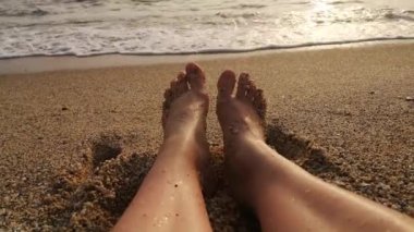 Gün batımının sıcak ışığında yavaşça hareket eden ayak parmaklarına yapışmış kaba kum taneleriyle ıslak kadın ayaklarının yakın çekimi. Sesli 4K video 15 saniye. Kumsalda yatarken birinci şahıs gözüyle bakıldı.