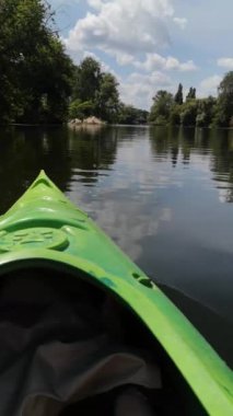 Yeşil kano, mavi bulutlu gökyüzünü yansıtan, bir nehrin ayna gibi sakin yüzeyinde yavaşça hareket ediyor. 4K dikey video 38 saniye. Nehir kıyısında kumlu bir sahil ve bereketli yaz yeşilliği yer alıyor.