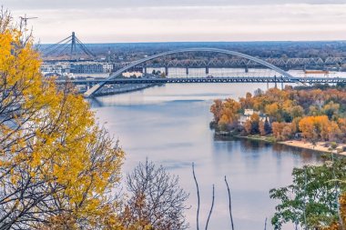 Ukrayna 'nın Kyiv kentindeki Dnipro Nehri ve Podilskyi Köprüsü' nün sonbahar yapraklarıyla çerçevelenmiş huzurlu bir manzarası. Nehrin sakin suları çevresindeki manzaranın güzelliğini ve modern köprü yapısını yansıtır.