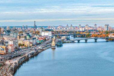 Ukrayna 'nın Kyiv kentinin panoramik manzarası, manzaralı Dnipro Nehri, hareketli sokaklar ve çeşitli mimari eserlerini sergiliyor. Görüntü, bulutlu bir gökyüzü altında modern binaların ve tarihi yapıların bir karışımını yakalıyor.