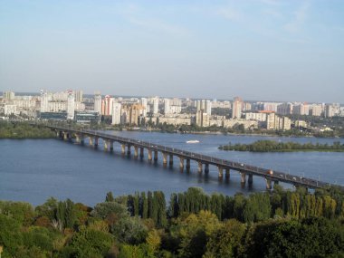 Kiev 'deki (Ukrayna) Dinyeper Nehri' nin karşısındaki Paton Köprüsü 'nün havadan görüntüsü. Mor-mavi gökyüzü, koyu mavi su yüzeyi, bir park ve nehrin karşı kıyısında kentsel bir bölge olan Kiev şehrinin güzel sulu bir manzarası.