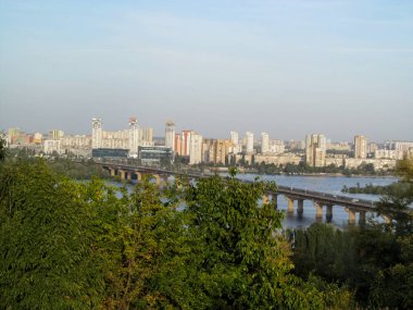 Anavatan Anıtı yakınlarındaki tepeden Kiev 'in manzarası. Ağaçların yeşil dallarının arkasında Paton Köprüsü 'nü, Dinyeper suyunun mavi yüzeyini ve nehrin diğer kıyısındaki Urban Bölgesi' ni görebilirsiniz.
