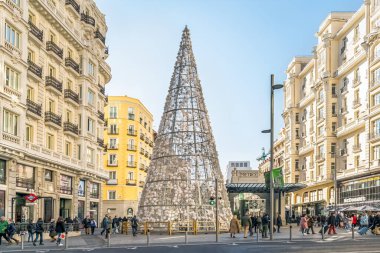 Madrid, İspanya - 30 Kasım 2021 Madrid 'de kalabalık bir Gran Via caddesinin ortasında tarihi binalar ve yayalarla çevrili büyük, konik bir Noel ağacı. Şehrin şenlikli atmosferi parlak güneş ışığıyla güçlenir.