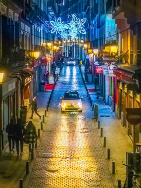 Madrid 'de Noel sezonunda kaldırım taşlı bir yolu aydınlatan şenlikli ışıkları gösteren bir gece sokak sahnesi. Sessiz caddede bir taksi sürüyor, tatil cazibesini artırıyor. Madrid, İspanya - 30 Kasım 2021