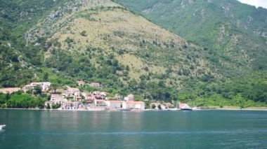 Karadağ 'ın Kotor kentinin dairesel panoramik manzarasında Kotor Körfezi' nin yeşil turkuaz sularında yelken açan bir tekne ve yat görülüyor. Video 27 saniye. Kıyı kasabaları dağların eteklerinde yuva yapar. Parlak bir yaz gökyüzünün altında.