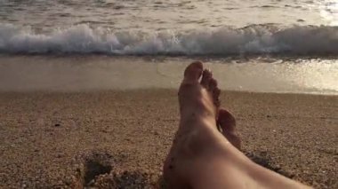 Altın gün batımında denizin yumuşak dalgaları tarafından çerçevelenmiş, kaba kumla kaplı bir sahilde dinlenen kadın ayaklarının birinci şahıs görüntüsü. 4K video 29 saniye. Sıcak, parlayan atmosfer dingin ve rahatlatıcı bir atmosfer yaratır.