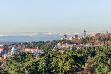 Madrid 'in panoramik manzarası. Yeşil ağaçların tepelerine ve arka plandaki binalara odaklanmış. Kubbeli bir yapı da dahil. Karlı tepeli uzak dağlar, kentsel ve doğal manzaraların karışımını gösteren manzaralı bir zemin sağlar.