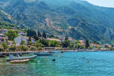 Kotor 'un Karadağ' daki kıyı şeridinin huzurlu bir manzarası, su boyunca kenetlenmiş tekneler ve yemyeşil dağların zeminine kurulmuş pitoresk evler. Sakin yaz atmosferi keşif ve rahatlamaya davetiye çıkarıyor.