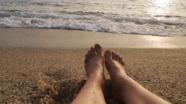 Batan güneşin altın ışığında deniz dalgaları kumlu bir sahili kaplar. Sesli 4K video, 30 saniye. Ön planda, çıplak ayaklı bir fotoğrafçı kumların üzerinde dinleniyor, birinci şahıs görüntüsünde yer alıyor.