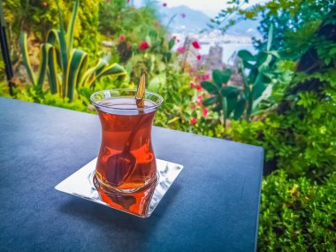 Bir bardak geleneksel Türk çayı, yemyeşil bitkiler ve canlı çiçeklerle çevrili açık bir masaya yerleştirilir. Huzurlu bahçe arkaplanı ferahlatıcı bir içeceğin tadını çıkarmak için huzurlu bir ortam yaratır.